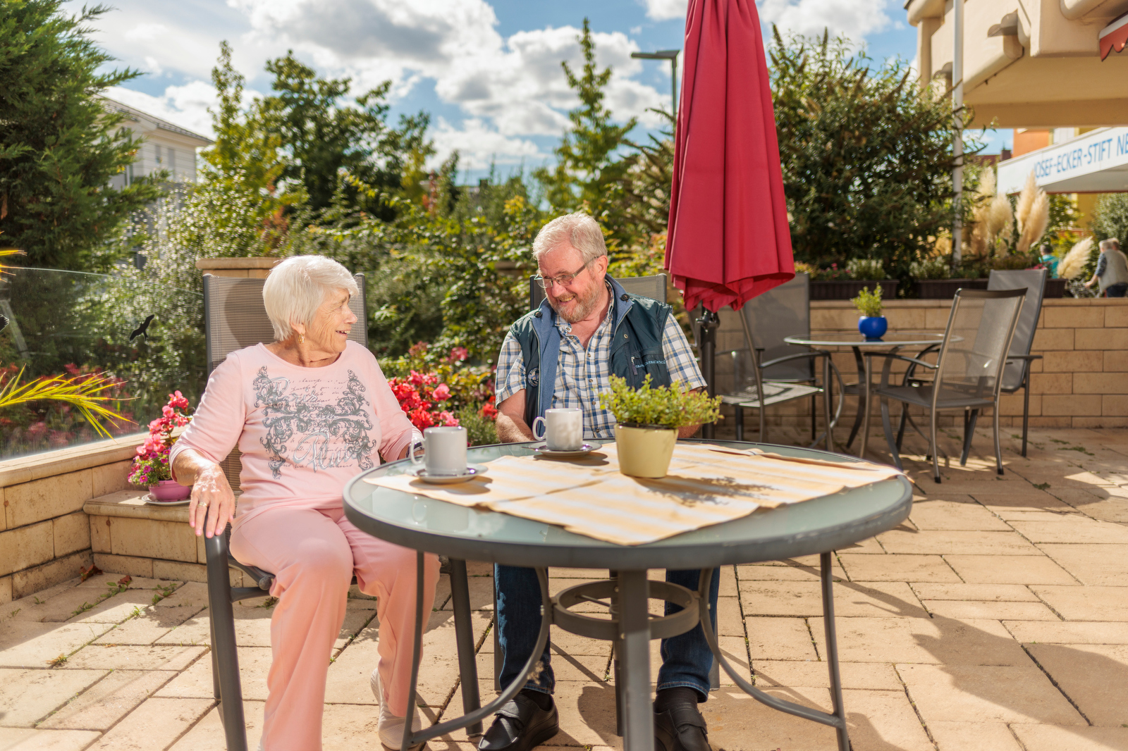 Zwei ältere Personen sitzen an einem runden Gartentisch mit Tischdecke, Tassen und einem kleinen Blumentopf, umgeben von Stühlen und Pflanzen auf einer Terrasse.