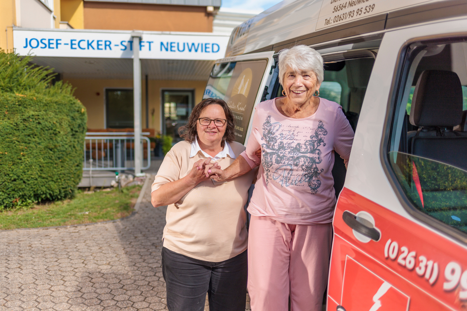 Zwei Frauen stehen nebeneinander vor einem roten Fahrzeug mit weißen Schriftzügen, im Hintergrund ein Gebäude mit dem Schriftzug 'JOSEF-ECKER-STIFT NEUWIED'.