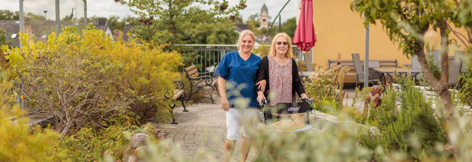Zwei Frauen gehen auf einem gepflasterten Weg in einem Garten, eine schiebt einen Rollator, die andere trägt eine blaue Bluse und weiße Hose.