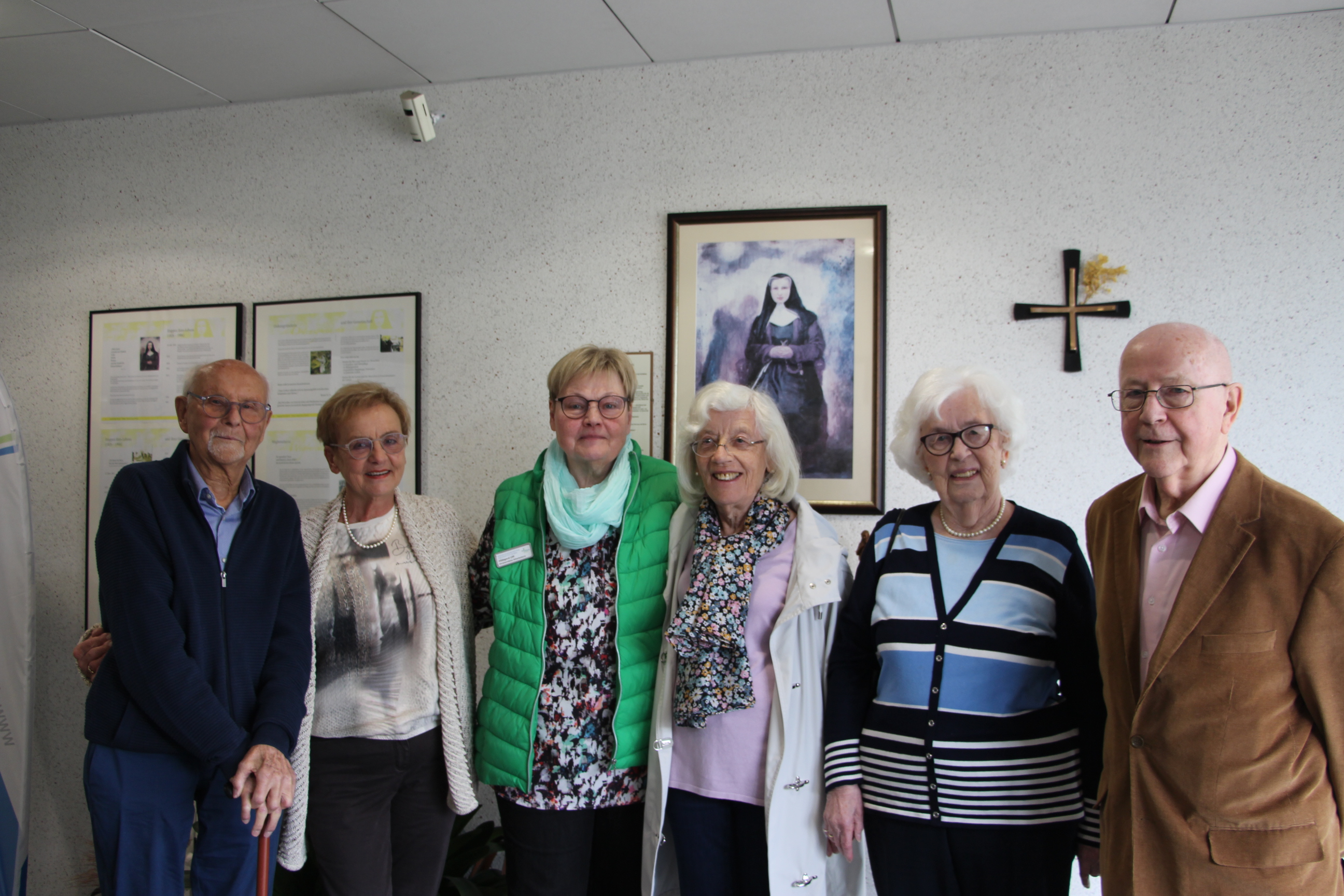 Sechs Personen stehen nebeneinander in einem Raum mit weißen Wänden, an der Wand hängen zwei gerahmte Dokumente und ein Bild einer Frau in dunkler Kleidung.