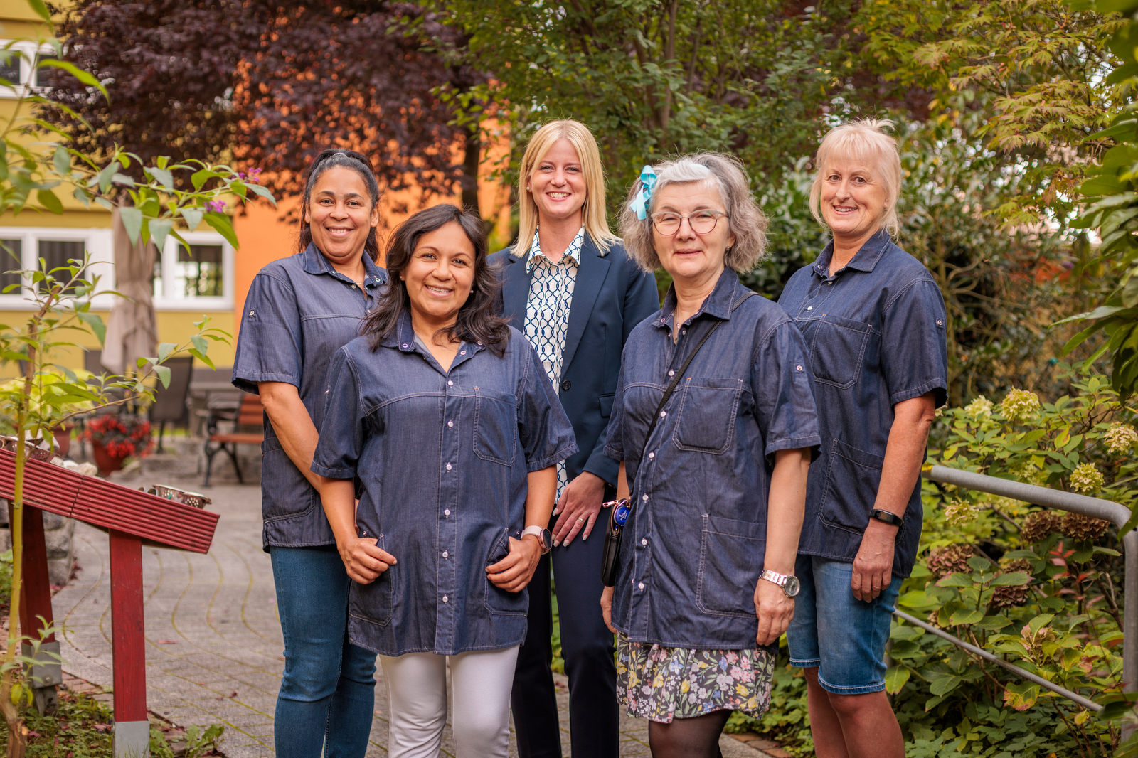 Fünf Frauen stehen in einem Gartenweg, vier tragen dunkelblaue Hemden, eine trägt ein dunkles Jackett und eine gemusterte Bluse.